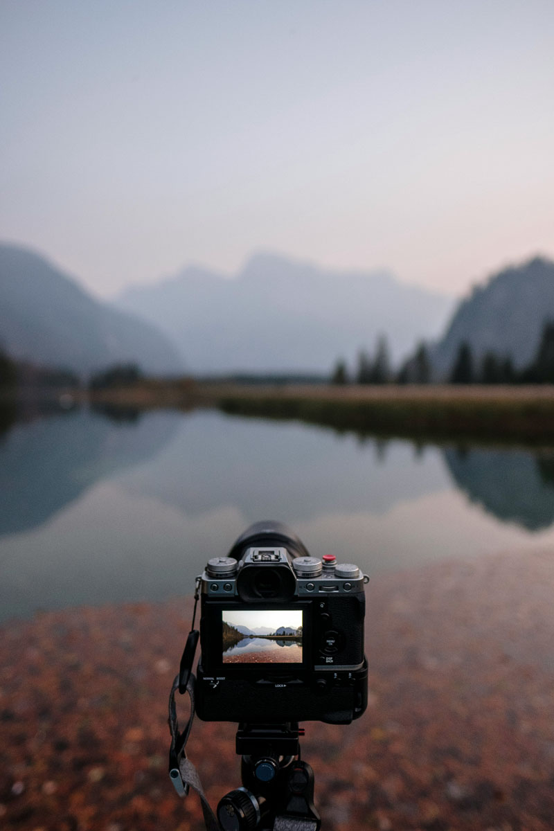 Camera on a tripod by a calm mountain lake at dusk.
